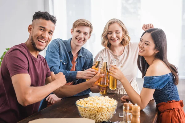 Joyful multicultural friends looking at camera while clinking bottles of beer — Stock Photo