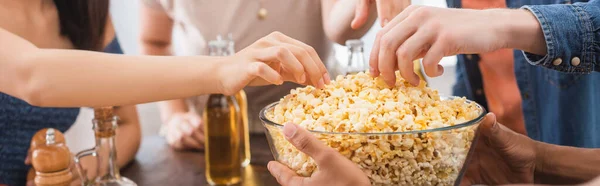 Selective focus of multicultural friends taking popcorn from bowl during party, horizontal crop — Stock Photo