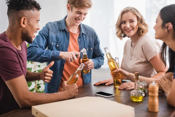Young man opening bottle of beer near multicultural friends during party — Stock Photo