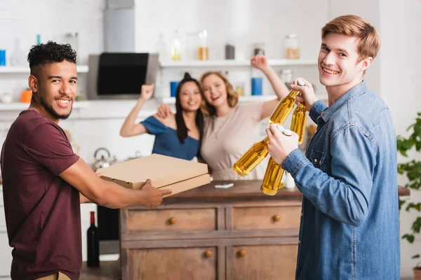 Selective focus of multicultural friends holding pizza boxes and beer near women showing yeah gesture — Stock Photo