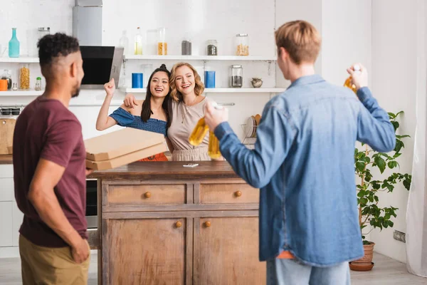 Vista trasera de amigos multiculturales sosteniendo cajas de pizza y cerveza cerca de mujeres mostrando sí gesto - foto de stock