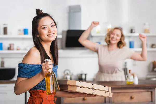 Foyer sélectif de la femme asiatique tenant des boîtes à pizza et de la bière près de l'ami sur fond — Photo de stock