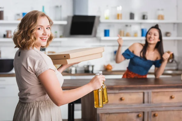 Enfoque selectivo de la mujer sosteniendo cajas de cerveza y pizza cerca de amigo asiático mostrando pulgares hacia arriba en el fondo - foto de stock