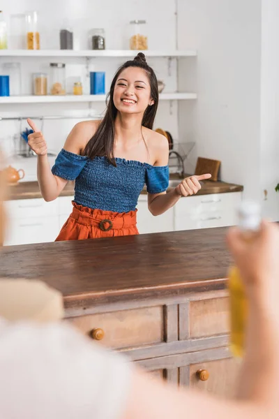 Selective focus of woman and joyful asian friend showing thumbs up in kitchen — Stock Photo