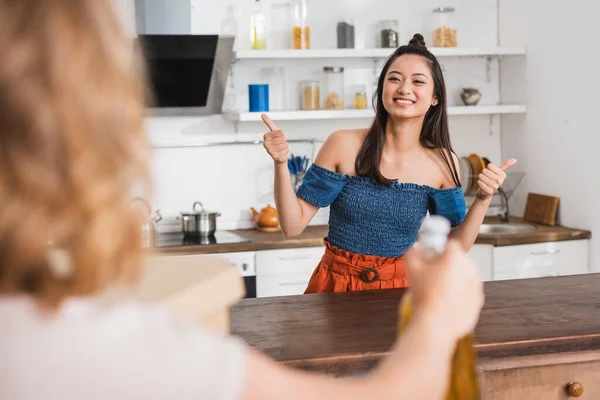 Selective focus of woman near excited asian friend showing thumbs up in kitchen — Stock Photo