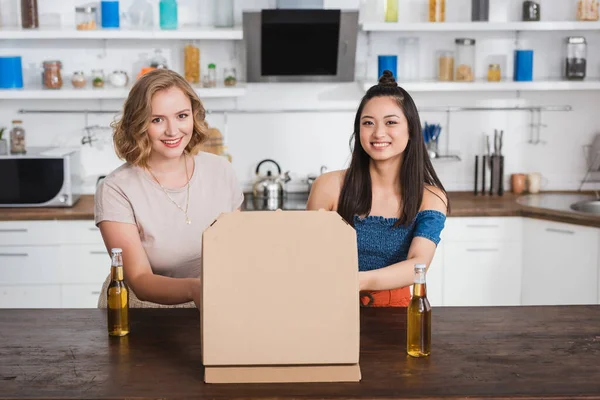 Joyful multicultural women looking at camera near beer and pizza box on table — Stock Photo