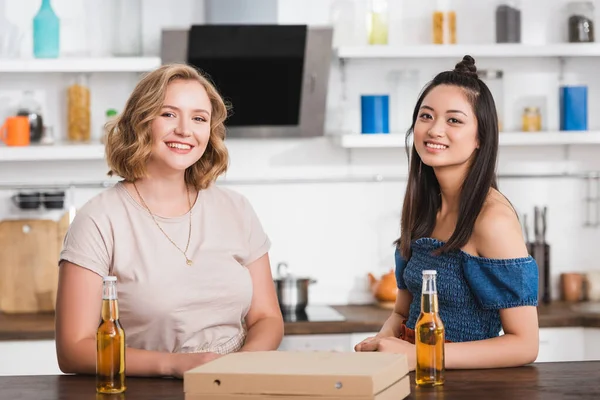 Молодые мультикультурные женщины смотрят в камеру рядом с пивом и коробкой пиццы на столе — стоковое фото