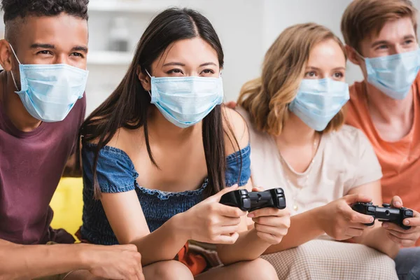 KYIV, UKRAINE - JULY 28, 2020: multicultural friends playing video game in medical masks during quarantine — Stock Photo