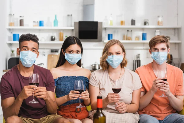 Multicultural friends in medical masks holding glasses of red wine while looking at camera in kitchen — Stock Photo