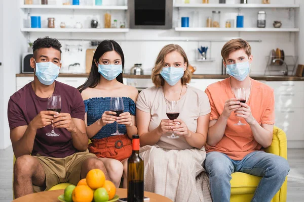 Multicultural friends in medical masks holding glasses of red wine on party during quarantine — Stock Photo