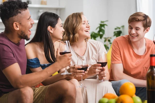 Amici multiculturali eccitati che parlano mentre tengono bicchieri di vino rosso in cucina — Foto stock