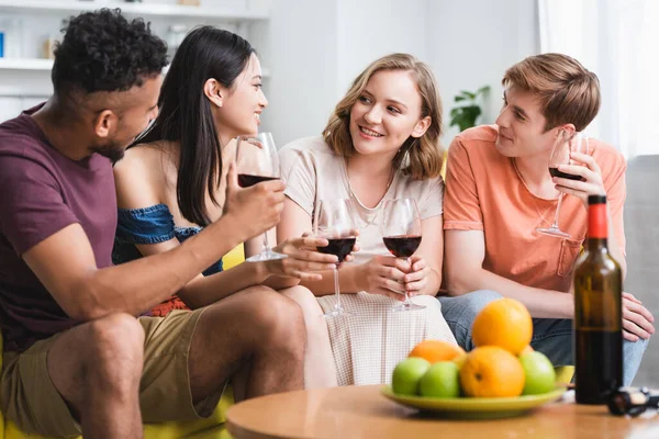 Multicultural friends talking while holding glasses of red wine in kitchen — Stock Photo