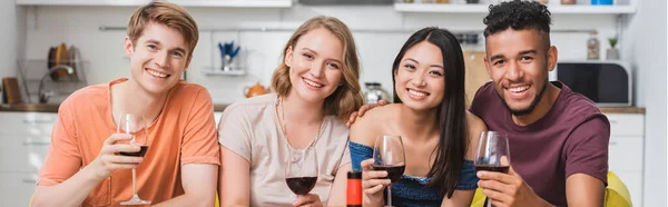 Horizontal image of multiethnic friends looking at camera while holding glasses of red wine in kitchen — Stock Photo