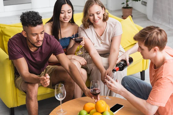 Visão de alto ângulo do jovem derramando vinho tinto perto de amigos multiculturais alegres e frutas frescas na cozinha — Fotografia de Stock