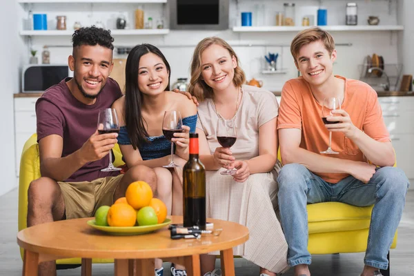 Multiethnic friends looking at camera while holding glasses of red wine in kitchen — Stock Photo