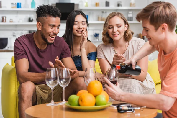Молодой человек наливает красное вино рядом с радостными мультикультурными друзьями и свежие фрукты на кухне — стоковое фото