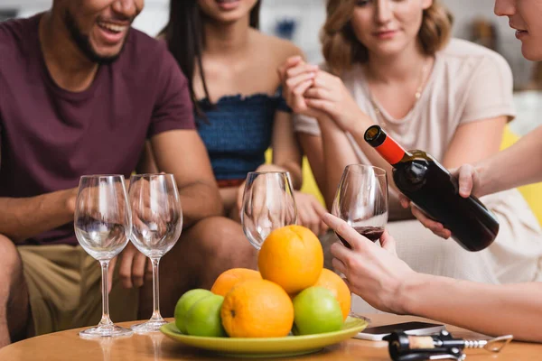 Cropped view of young man pouring red wine near fruits and multicultural friends — Stock Photo
