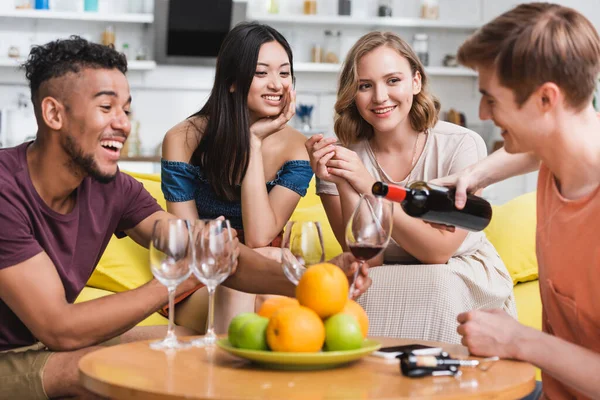 Giovane versando vino rosso vicino ad amici multietnici e frutta fresca in cucina — Foto stock