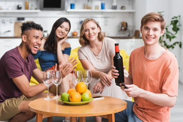 Giovane in possesso di bottiglia di vino rosso vicino amici multiculturali e frutta fresca in cucina — Foto stock