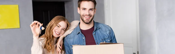 Plano panorámico de mujer alegre sosteniendo llaves cerca de novio con caja de cartón, concepto en movimiento - foto de stock