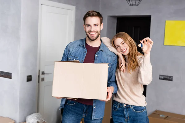 Mujer alegre sosteniendo llaves cerca de novio con caja de cartón — Stock Photo