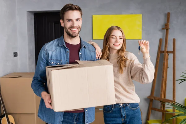 Mujer alegre mirando las llaves cerca de novio sosteniendo caja de cartón — Stock Photo