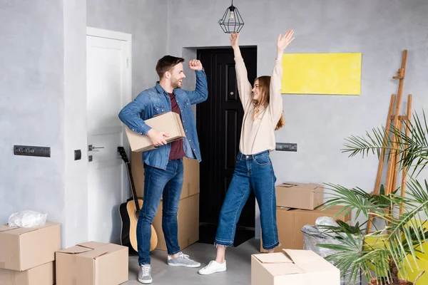 Homme excité tenant boîte en carton et regardant femme heureuse debout avec les mains au-dessus de la tête, concept de relocalisation — Photo de stock