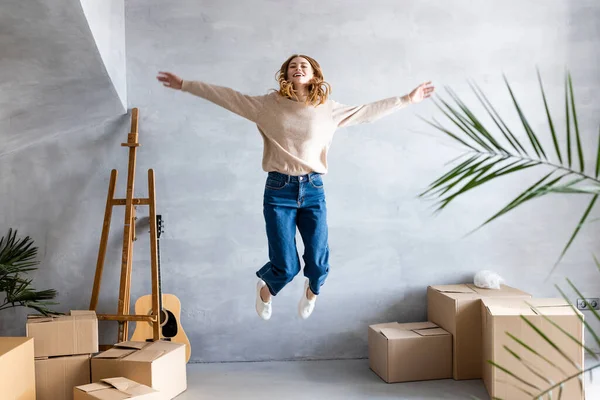 Young pleased woman with outstretched hands jumping near carton boxes, relocation concept — Stock Photo