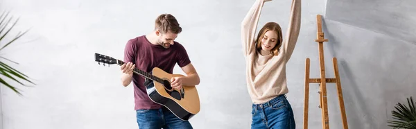 Plano panorámico del hombre tocando la guitarra acústica cerca de mujer complacida bailando con las manos por encima de la cabeza - foto de stock