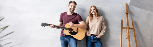 Website header of man playing acoustic guitar near pleased woman standing with hands in pockets near easel — Stock Photo