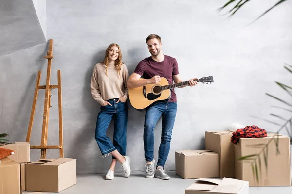 Foco seletivo do homem tocando guitarra acústica perto da mulher satisfeita de pé com as mãos nos bolsos perto de cavalete e caixas — Fotografia de Stock