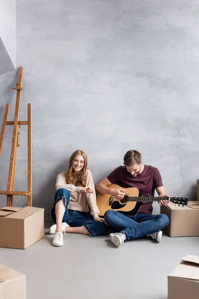 Hombre tocando la guitarra acústica cerca de la mujer complacida sentado cerca del caballete y cajas, concepto en movimiento - foto de stock