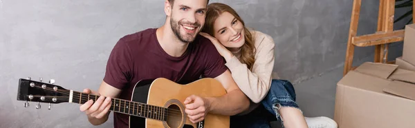 Imagen horizontal de la mujer complacida abrazando novio tocando la guitarra acústica cerca de cajas, concepto de reubicación - foto de stock