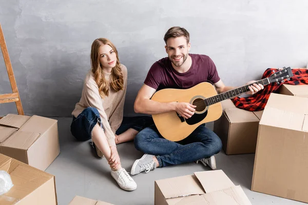 Молода жінка сидить на підлозі з чоловіком грає на акустичній гітарі біля коробки, концепція переміщення — стокове фото