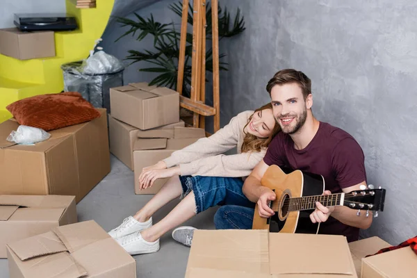 Радостная женщина опирается на бойфренда, играющего на акустической гитаре и сидящего на полу рядом с коробками, концепция переезда — стоковое фото