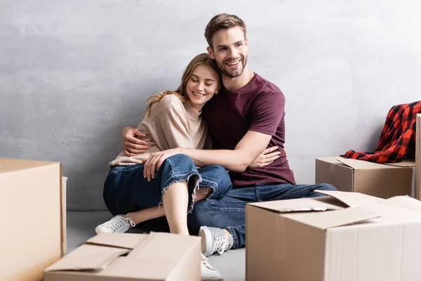 Hombre complacido sentado de piso y abrazando a mujer cerca de cajas — Stock Photo