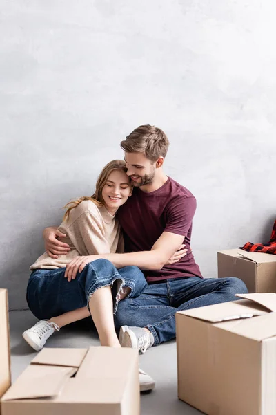 Zufriedener Mann sitzt auf dem Boden und umarmt freudige Freundin in der Nähe von Boxen — Stockfoto