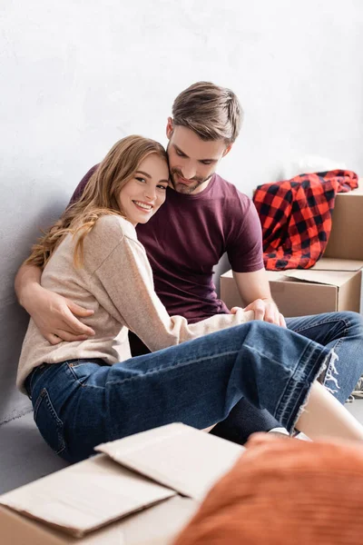 Complacido hombre sentado de piso y abrazando novia cerca de cajas - foto de stock