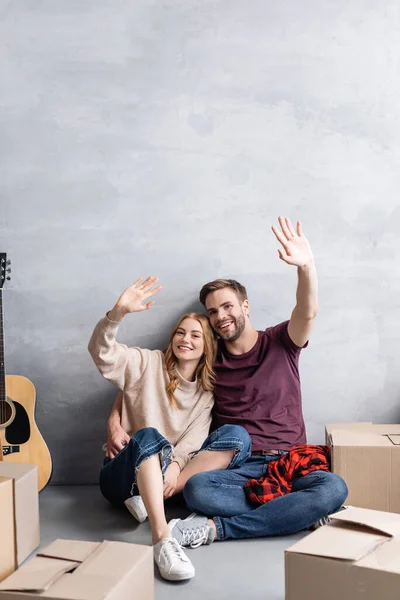 Couple waving hands and sitting near acoustic guitar and boxes — Stock Photo