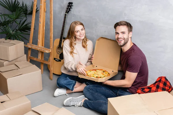 Joyful couple looking at camera while holding pizza near carton boxes and acoustic guitar, moving concept — Stock Photo