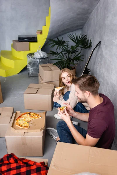 Femme à bouche ouverte regardant petit ami avec pizza dans les mains près des boîtes en carton — Photo de stock