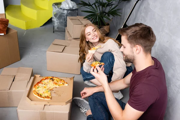 Selektiver Fokus der fröhlichen Frau, die Mann mit Pizza in der Nähe von Pappkartons auf dem Boden betrachtet — Stockfoto