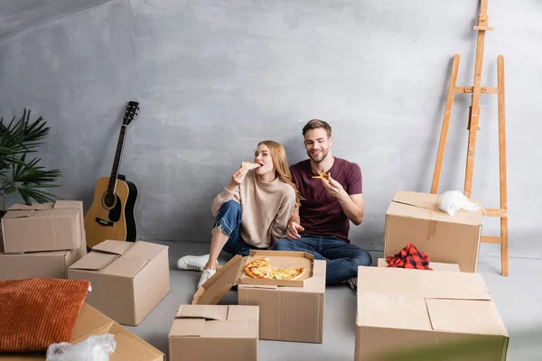 Mujer joven comiendo pizza cerca de hombre y cajas de cartón, concepto de reubicación - foto de stock