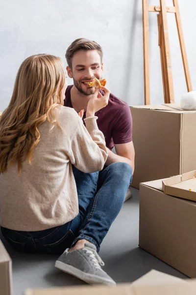 Young woman feeding pleased boyfriend with pizza near boxes — Stock Photo