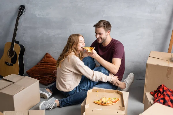 Pleased man feeding girlfriend with pizza near carton boxes — Stock Photo