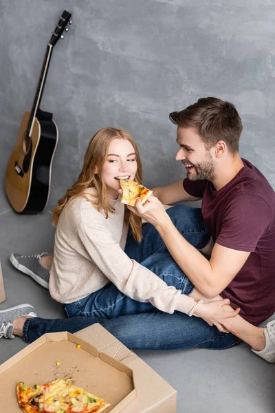 Homem alegre alimentando namorada com pizza perto de caixas de papelão, conceito em movimento — Fotografia de Stock