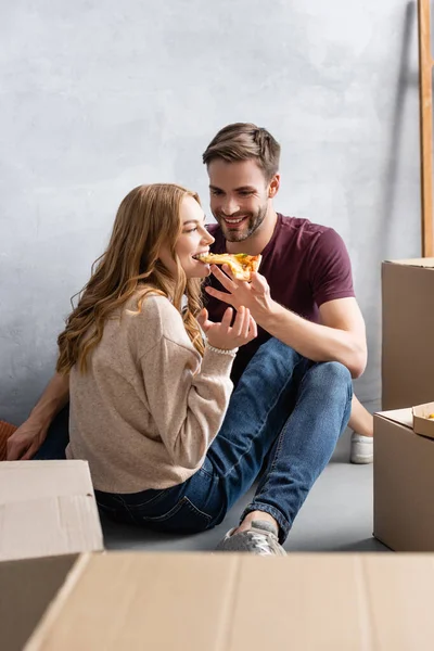 Selective focus of joyful man feeding pleased girlfriend with pizza near carton boxes, moving concept — Stock Photo