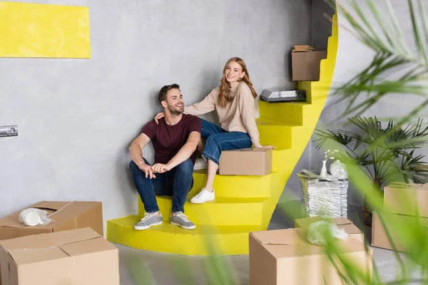 Selektiver Fokus des fröhlichen Paares, das auf gelben Treppen in der Nähe von Kartons sitzt, bewegtes Konzept — Stockfoto
