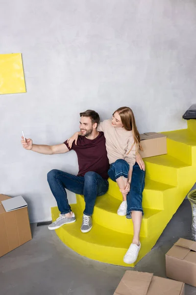 Heureux jeune couple assis sur des escaliers jaunes et prenant selfie près des boîtes en carton — Photo de stock