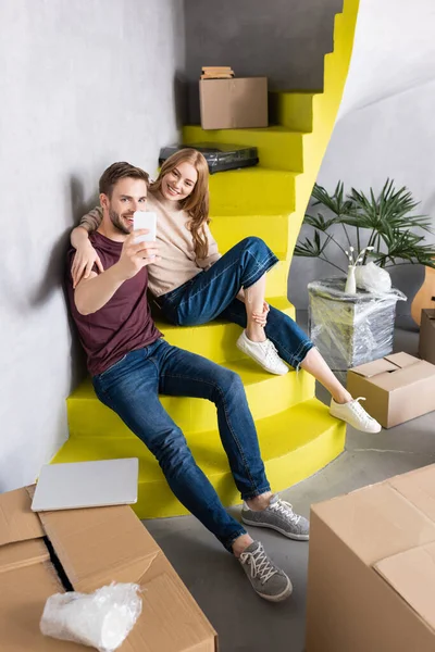 Heureux couple assis sur les escaliers jaunes et prendre selfie près des boîtes en carton — Photo de stock